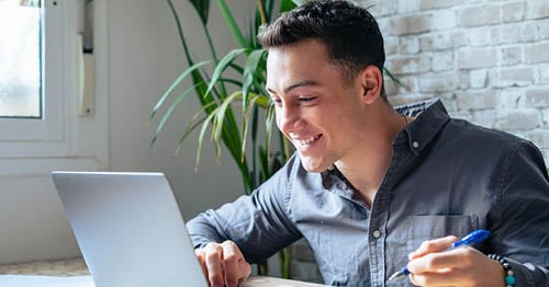  An online student in a blue shirto holding a pen sitting at a desk working on an online assignment for Connections Academy. 